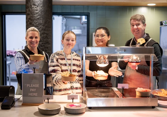 Four people stand behind a bain marie and cafe counter holding up small bowls with rice and curry