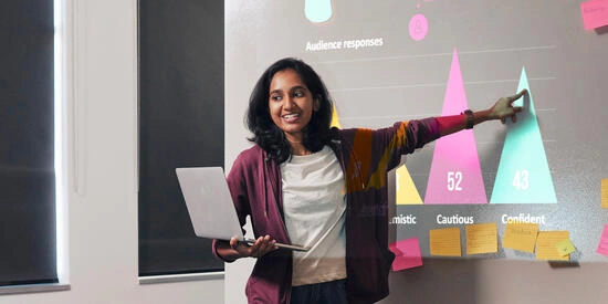 A student holding a laptop and pointing at a slide they're presenting on. 