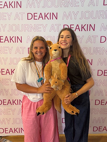 Zoe and a peer in front of a Deakin banner, holding a toy kangaroo and joey