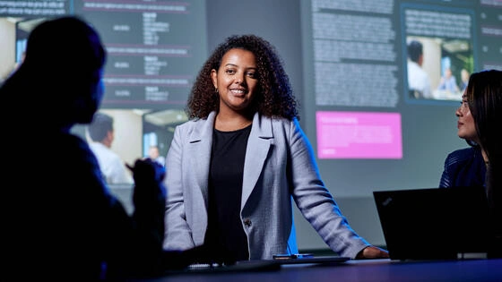 Three Deakin business students inside classroom with data graphs on a presentation screen.