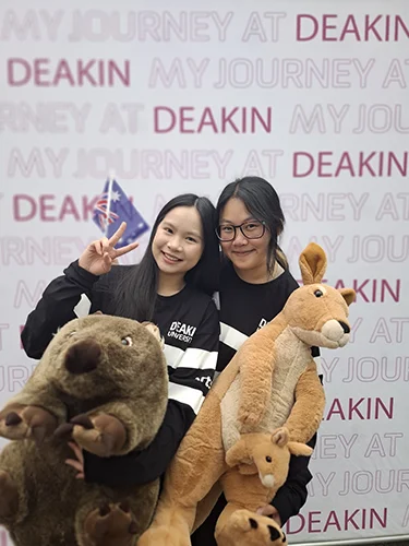 Joycely 3 Joycely and a friend in front of a Deakin banner, holding stuffed toys