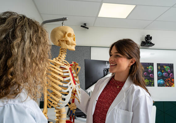 A medical student wearing a white coat examining a skeleton.