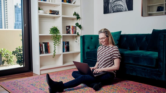 Postgraduate student smiling and sitting at home on the floor with a laptop resting on her.